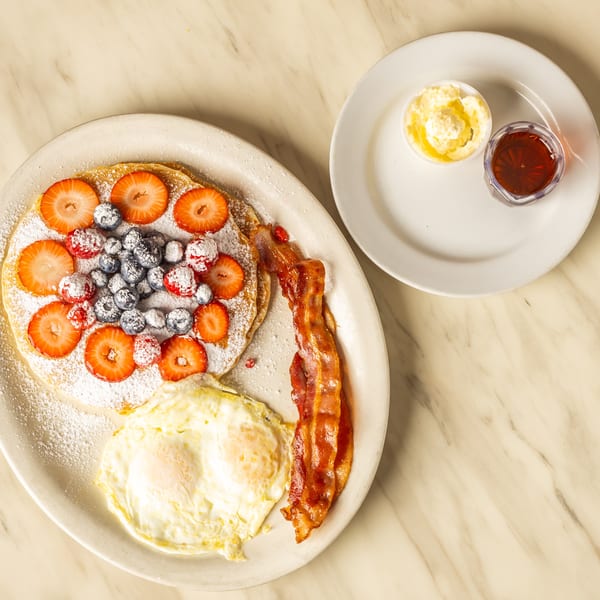 Pancakes with berries, powdered sugar, bacon, and fried eggs, with syrup and butter on the side.