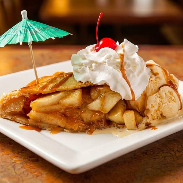 Caramel apple pie slice with vanilla ice cream, whipped cream, cherry, and a green umbrella.