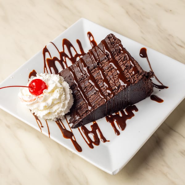 A slice of chocolate cake with chocolate drizzle, whipped cream, and a cherry on a white plate.