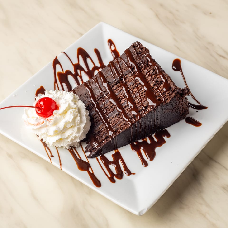 A slice of chocolate cake with chocolate drizzle, whipped cream, and a cherry on a white plate.