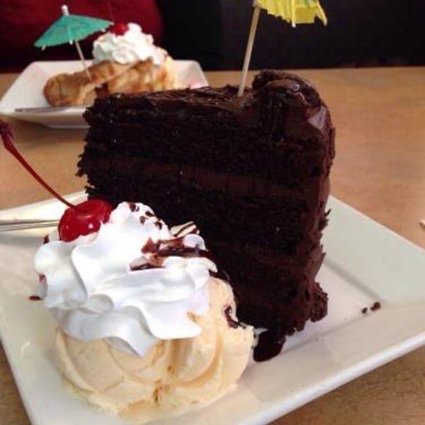 Slice of chocolate cake with ice cream, whipped cream, cherry, and yellow umbrella, another dessert in background.