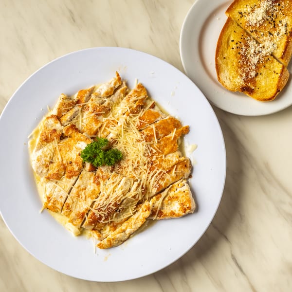 Sliced chicken breast with creamy sauce, shredded cheese, and parsley, served with garlic bread.