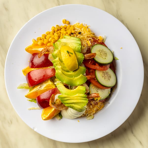 Colorful salad with shrimp, avocado, corn, tomatoes, cucumbers, and fruit on a white plate.
