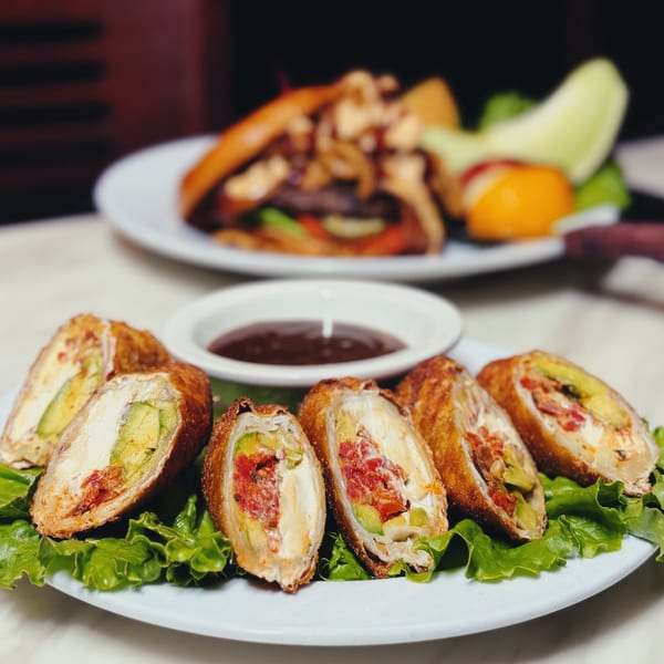 Platter of sliced golden appetizers stuffed with vegetables on lettuce, with dipping sauce and a grilled entrée in the background.